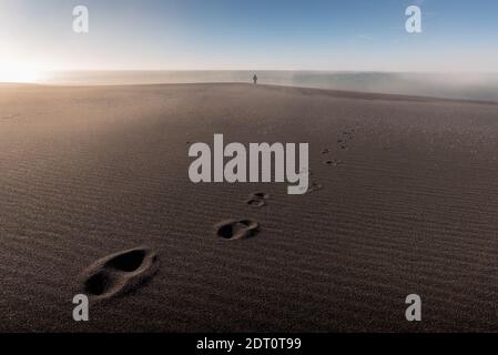 Einsame Person Silhouette steht weit im Hintergrund und Fußabdrücke auf Der Sand im Vordergrund Stockfoto