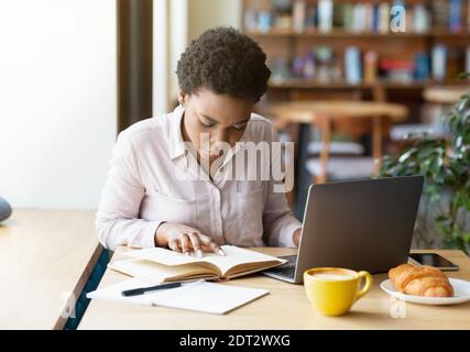 Smart schwarze Frau Buch vor dem Laptop im Café lesen, sich bereit für die Prüfung oder die Arbeit an Business-Projekt Stockfoto
