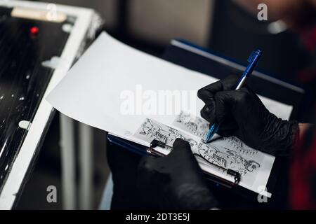 Tattoo-Künstler im Salon zeichnet eine schöne Skizze eines Stier-Tattoo auf Papier mit einem Stift für seinen Kunden. Nahaufnahme Stockfoto