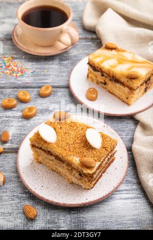 Honigkuchen mit Milchcreme, Karamell, Mandeln und einer Tasse Kaffee auf grauem Holzhintergrund und Leinentextilien. Seitenansicht, Nahaufnahme. Stockfoto