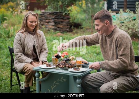 Ein Mann legt Teller mit gebratenem Fleisch auf den Picknicktisch. Die letzten warmen Herbsttage vor dem Winter. Gemütliches Picknick im Hinterhof eines privaten Hauses Stockfoto