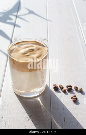 Traditionelles koreanisches Getränk, Dalgona Kaffee in einem transparenten Glas mit Kaffeebohnen auf einem Hintergrund von weißen Brettern. Stockfoto