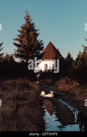 Kleine Kapelle mit Bergweg mit Pfütze bei Sonnenuntergang Stockfoto