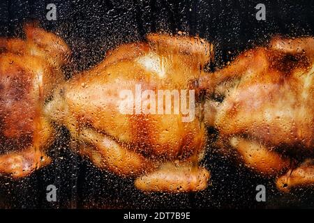 Nahaufnahme eines dampfenden Maschinenfensters mit Wassertropfen. Gegrillte gebratene ganze Hühner mit schmackhafter goldgelber gebratener Haut auf einem Spieß. Stockfoto