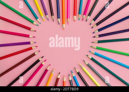 Herz mit geschärften mehrfarbigen Bleistiften auf einem rosa Hintergrund ausgelegt, flach legen, kopieren Raum. Stockfoto