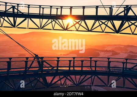 Silhouette der Brücke des Aussichtsturms Stezka V oblacích in Tschechien mit Blick auf schneebedeckte Hügel und Bäume bei farbenfrohem Sonnenuntergang. Stockfoto