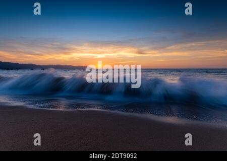 Ein farbenfroher Sonnenuntergang am Meer mit einer Welle, die an der Küste bricht Die Sonne untergeht am Horizont Stockfoto