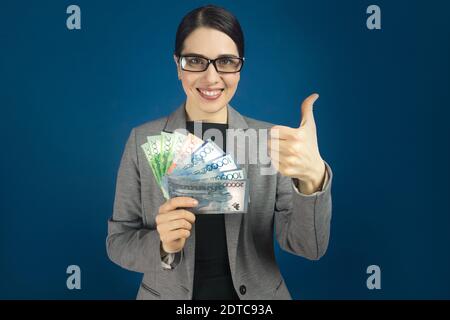 Glücklich lächelnde junge Frau in einer Brille mit Tenge in den Händen, zeigt Daumen nach oben. Stockfoto