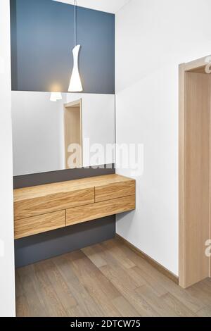 Stilvoller Schrank aus hellbraunem Naturholz an der Wand montiert Unter großem Spiegel und glühender Lampe im modernen Apartment-Interieur Design Stockfoto