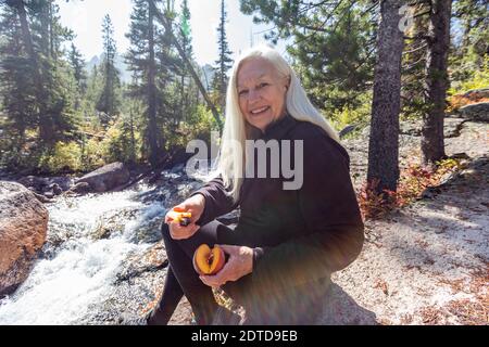 USA, Idaho, Stanley, ältere weibliche Wanderer lächeln und schneiden frischen Pfirsich neben Strom Stockfoto