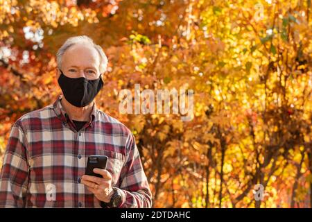 Senior Mann trägt Covid Schutzmaske und hält Smartphone im Freien Im Herbst Stockfoto