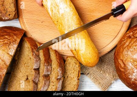 Ein französisches Baguette auf einem hellen Tisch schneiden. Brotscheiben, weißes Baguette und eine Frauenhand mit einem Messer. Blick von oben. Stockfoto