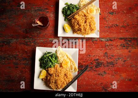 Flaches Lay von gebratenen Wontonnudeln mit gebratenem Ei Und Spinat gegen einen rustikalen roten Tisch Stockfoto