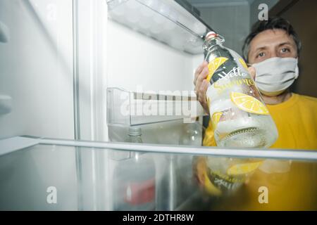 Mann mittleren Alters, der eine Maske mit einer Glasflasche trägt An die Kamera, während sie in der Nähe der offenen Tür von steht Der leere Kühlschrank Stockfoto
