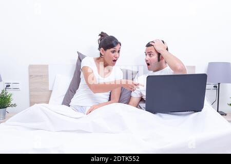 Glückliches junges Paar mit Laptop im Bett zu Hause, sie scheinen sehr überrascht. Intermet und Arbeit zu Hause Konzept . Stockfoto