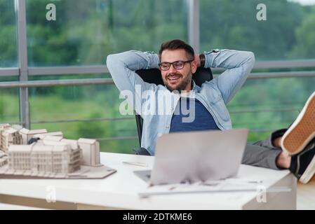 Zufriedener bärtiger männlicher Designer in Brille sitzend in entspannter Pose im Designstudio zufrieden mit seinem Projekt. Stockfoto