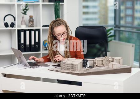 Erfahrene stilvolle Architektin mit Dreadlocks arbeiten mit Pappe Modell Von Wohngebiet in Architekturbüro Stockfoto