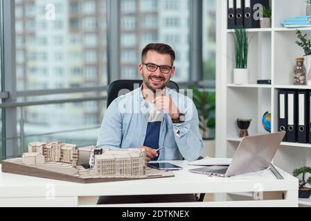 Sympathisch erfolgreicher bärtiger 35-Jähriger Architekt in Brille sitzt an seinem Arbeitsplatz mit einem Modell zukünftiger Gebäude. Stockfoto