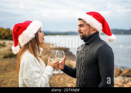 Schöne heterosexuelle Paar in der Liebe Toasten im Freien trägt Santa Claus hut in die Augen schauen, hält Weißweinglas In der Hand mit se Stockfoto