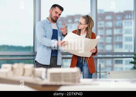 Selbstbewusster bärtiger männlicher Designer und seine Kollegin mit Dreadlocks Analyse gedruckt auf Papier-Projekt. Stockfoto