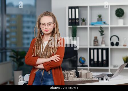 Stilvolle Architektin in Gläsern mit Dreadlocks sitzt an ihrem Arbeitsplatz mit Mock-up des zukünftigen Gebäudes. Stockfoto