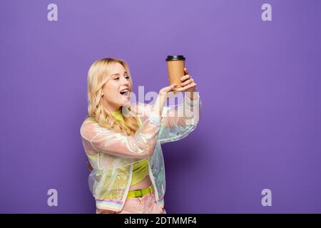 Aufgeregt blonde junge Frau in bunten Outfit mit Papierbecher Auf violettem Hintergrund Stockfoto