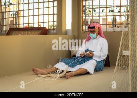 Arabischer Fischer, Reparatur von hausgemachten Fischernetz und Holzyacht, trägt Gesichtsmaske und sitzt auf dem Boden der traditionellen Boam Stockfoto