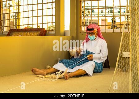 Arabischer Fischer, Reparatur von hausgemachten Fischernetz und Holzyacht, trägt Gesichtsmaske und sitzt auf dem Boden der traditionellen Boam Stockfoto