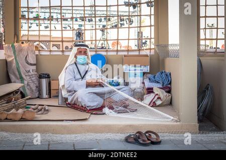 Arabischer Fischer, Reparatur von hausgemachten Fischernetz und Holzyacht, trägt Gesichtsmaske und sitzt auf dem Boden der traditionellen Boam Stockfoto
