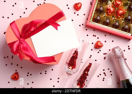 Herzförmige Schachtel mit leerer Papierkarte, Schachtel Pralinen, Flasche Champagner und Gläsern mit Konfetti auf rosa Hintergrund. Valentinstag Abendessen, l Stockfoto