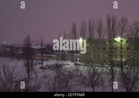 In der Winternacht werden Autos auf dem Parkplatz, um Bäume, Gebäude, Häuser geparkt. Alles ist mit Schnee bedeckt Stockfoto