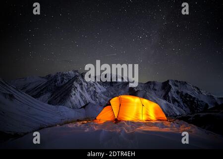 Leuchtendes orangefarbenes Zelt in den Winterbergen bei dunkler Nacht Himmel Hintergrund mit Sternschnuppe