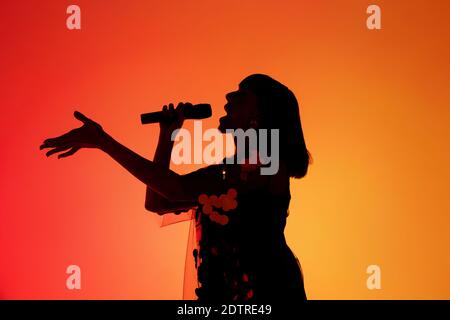 Talentiert. Silhouette der jungen Sängerin isoliert auf orange Gradient Studio Hintergrund in Neonlicht. Schöner Schatten in Aktion, Leistung. Konzept der menschlichen Emotionen, Ausdruck, Anzeige, Musik, Kunst. Stockfoto