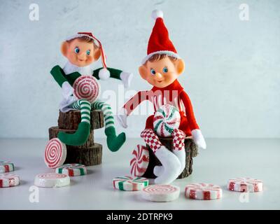 Zwei Weihnachtselfen mit roten und grünen Outfits sitzen auf Holz, halten große Stücke von Süßigkeiten mit mehr Süßigkeiten verstreut herum. Weihnachtsdekor, Weihnachtsmann Stockfoto