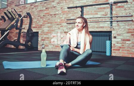 Fröhliche sportliche Frau sitzt und ruht sich auf einer Trainingsmatte aus Und schreibt zukünftige Trainingspläne auf, um hervorragende Ergebnisse zu erzielen Im Fitnessstudio Stockfoto