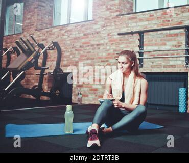 Fröhliche sportliche Frau sitzt auf einer Trainingsmatte und schreibt Down zukünftige Trainingspläne für die Erreichung großer Ergebnisse im Fitnessstudio Stockfoto