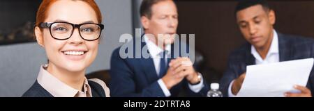 Glücklich Rotschopf Geschäftsfrau Blick auf Kamera mit verschwommenen multikulturellen Kollegen auf Hintergrund, Banner Stockfoto