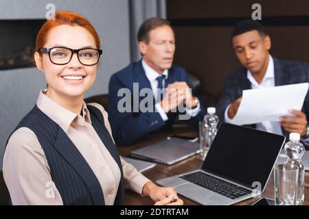 Lächelnde Geschäftsfrau, die die Kamera anschaut, während sie am Arbeitsplatz mit sitzt Digitale Geräte und verschwommene Kollegen im Hintergrund Stockfoto