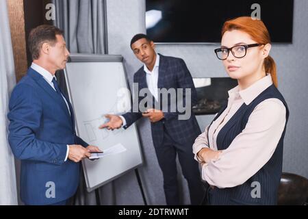Selbstbewusster Rotschopf-Manager, der mit verschwommenen multikulturellen Geschäftsleuten in die Kamera schaut Gestikulieren auf dem Hintergrund Stockfoto