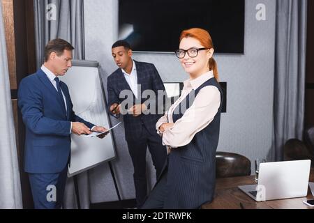 Lächelnd Rotschopf Geschäftsfrau mit gekreuzten Armen Blick auf Kamera mit Multikulturelle Kollegen im Hintergrund Stockfoto