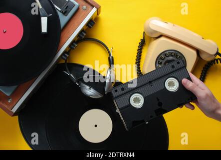 Weibliche Hand hält die Videokassette. 80er-Jahre-Style. Vinyl-Player, Kopfhörer, Drehtelefon auf gelbem Hintergrund. Draufsicht, flach liegend Stockfoto