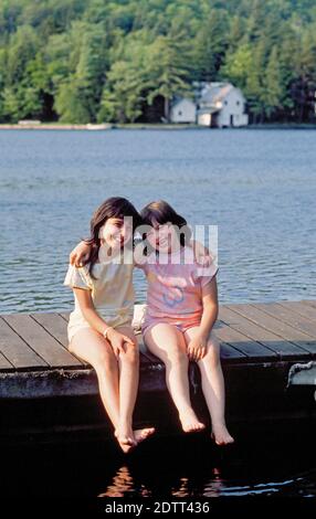 Zwei junge Mädchen im Urlaub mit ihren Eltern sitzen darauf Ein Holzdock an einem Erholungsgebiet am Finger Seen in den Adirondacks im Bundesstaat New York Stockfoto