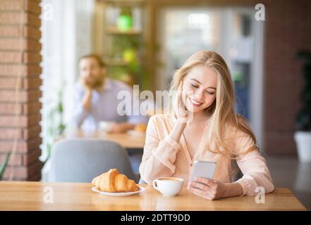 Schöne junge Frau mit Smartphone während des Frühstücks im Café, junge Mann, der sie anschaut, leerer Raum Stockfoto
