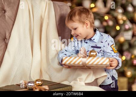 Ein schöner Junge von 4-5 Jahren steht in der Nähe des Sofas und hält Geschenke in seinen Händen. Das Konzept der Winterferien. Selektiver Weichfokus, Bokeh (Unschärfe Stockfoto