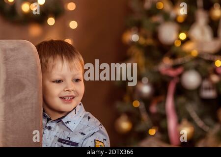 Ein schöner Junge von 4-5 Jahren steht (sitzt) in der Nähe des Sofas und träumt von einem Urlaub und Geschenke. Das Konzept der Winterferien. Selektiver Weichfokus, Stockfoto