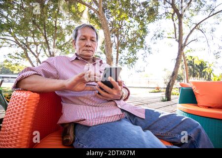 Asiatische ältere Menschen mit grauen Haaren verwenden Mobiltelefone. Ein glücklicher älterer Mann, der auf einem bequemen Sofa im Garten sitzt. Stockfoto