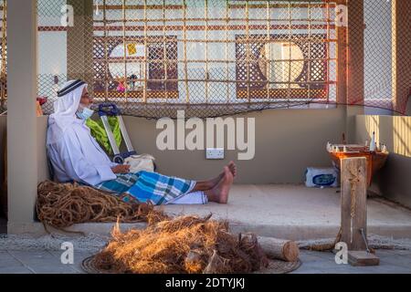 Arabischer Fischer, Reparatur von hausgemachten Fischernetz und Holzyacht, trägt Gesichtsmaske und sitzt auf dem Boden der traditionellen Boam Stockfoto