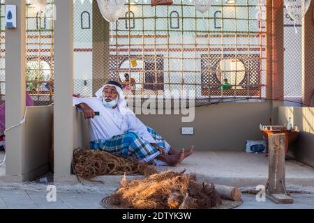 Arabischer Fischer, Reparatur von hausgemachten Fischernetz und Holzyacht, trägt Gesichtsmaske und sitzt auf dem Boden der traditionellen Boam Stockfoto