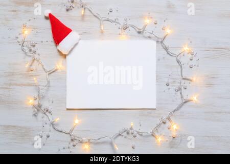 Grußkarte auf Vintage Holz Hintergrund mit Weihnachtsmann Hut und glänzenden Girlande Lichter. Leere Grußkarte mit Weihnachtsbeleuchtung. Minimale Weihnachten Stockfoto