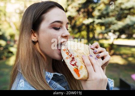 Ein hungriges Mädchen bisste ein Sandwich im Park und Bedeckte ihre Augen mit Vergnügen Stockfoto
